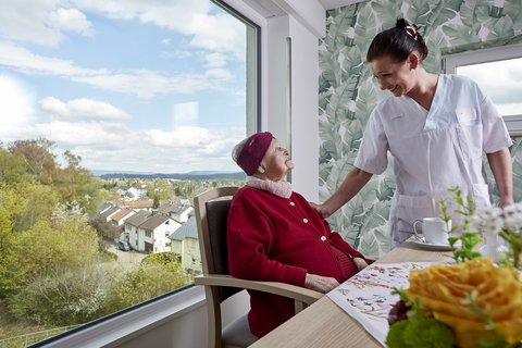 Bewohnerin sitzt im Aufenthaltsraum und sieht Pflegekraft an