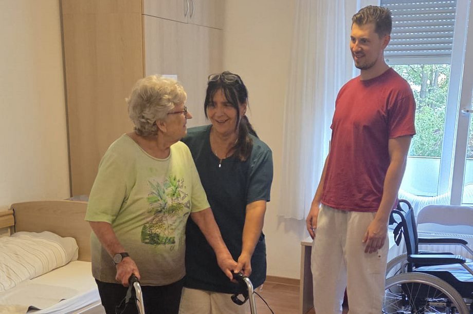 Eine ältere Person nutzt in einem Pflegezimmer einen Rollator. Zwei Pflegekräfte begleiten die Übung.