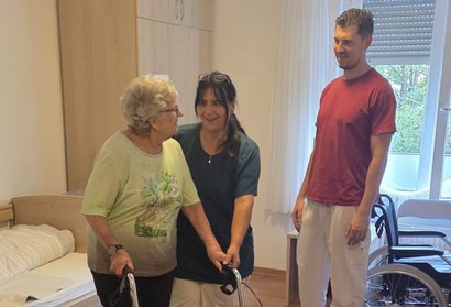 Eine ältere Person nutzt in einem Pflegezimmer einen Rollator. Zwei Pflegekräfte begleiten die Übung.