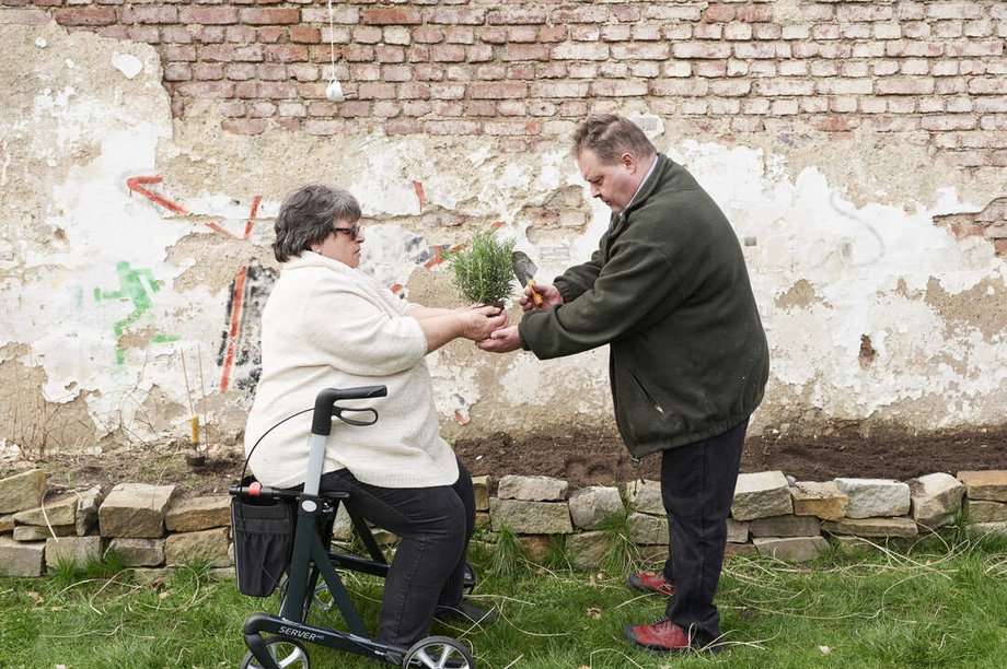 Mann überreicht Pflanze an eine auf Rollator sitzende Seniorin.