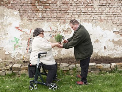 Mann überreicht Pflanze an eine auf Rollator sitzende Seniorin.